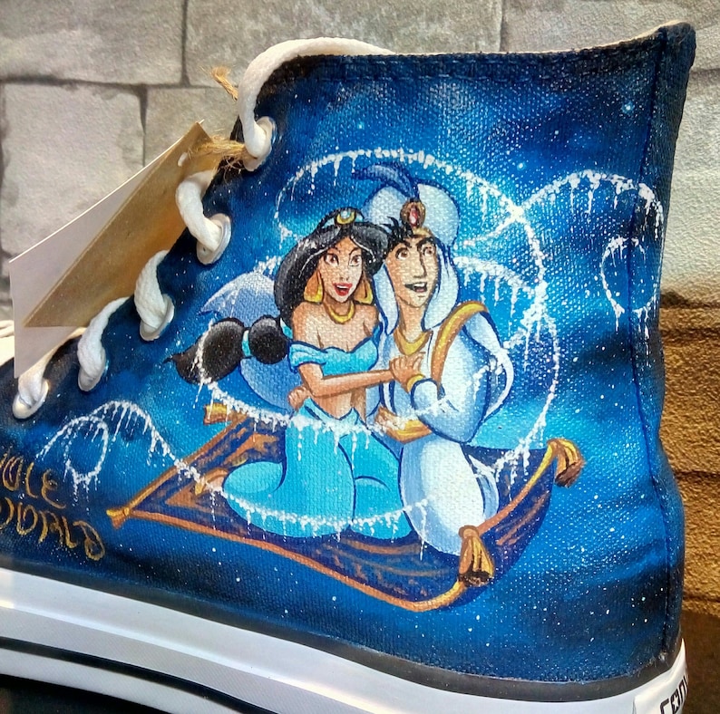 Op de afbeelding: Hoge canvas schoen met een handgeschilderd ontwerp van Aladdin en Jasmine op een vliegend tapijt tegen een sterrenblauwe achtergrond. De schoen heeft witte veters en een witte zool. De woorden "WHOLE WORLD" zijn zichtbaar.