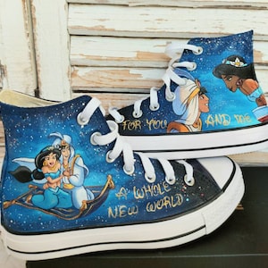 Op de afbeelding: Op maat geschilderde high-top sneakers met een ontwerp van een sterrenhemel. De schoenen tonen personages uit Aladdin, met de tekst "A Whole New World" en "For You And Me" in gouden letters. Witte veters en zolen.