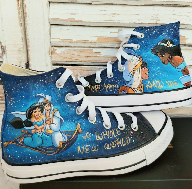 Op de afbeelding: Op maat geschilderde high-top sneakers met een ontwerp van een sterrenhemel. De schoenen tonen personages uit Aladdin, met de tekst "A Whole New World" en "For You And Me" in gouden letters. Witte veters en zolen.