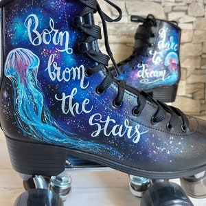 May include: A pair of black roller skates with a galaxy-themed design. The skates feature a blue and purple galaxy background with white stars and a jellyfish. The left skate has the text "Born from the Stars" written in white cursive. The right skate has the text "Dare to Dream" written in white cursive.