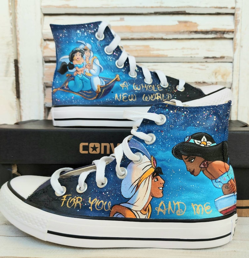 Op de afbeelding: Op maat geschilderde hoge sneakers met Aladdin en Jasmine uit Disney's Aladdin. De schoenen hebben een blauwe sterrennacht achtergrond met de tekst "A Whole New World" en "For You And Me". De sneakers hebben witte veters en zolen.