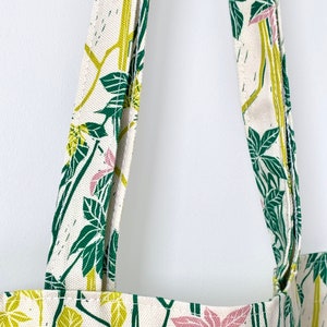 May include: Close-up of the straps of a white tote bag with a green and pink floral pattern. The straps are made of the same fabric as the bag.