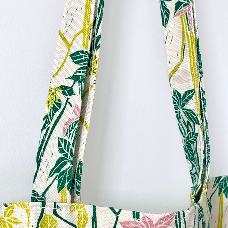 May include: Close-up of the straps of a white tote bag with a green and pink floral pattern. The straps are made of the same fabric as the bag.