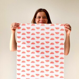 May include: A rectangular white canvas with a repeating pattern of red and yellow watermelon slices. The canvas is held up by a person. The watermelon slices are arranged in rows and columns.