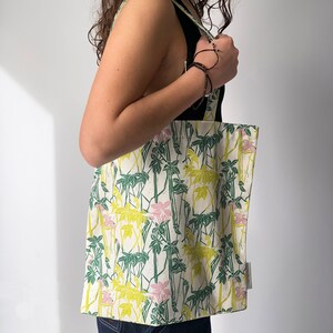 May include: A white tote bag with a floral print featuring green leaves, pink and yellow flowers, and white stems. The bag has a black strap and is being held by a person.