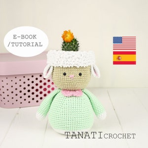 May include: A crocheted sheep planter with a green body, white head, and pink collar. A small cactus with orange flowers is planted in the sheep's head. The planter is sitting on a white wooden surface. E-BOOK / TUTORIAL is written in a white circle on the left side of the image. The American and Spanish flags are in the upper right corner of the image. TANATI CROCHET is written in white letters on the bottom right corner of the image.