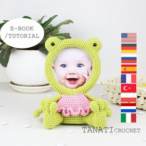 Puede incluir: Un marco de fotos de rana de crochet verde con un vestido de crochet rosa. El marco contiene una foto de un bebé sonriendo. El texto "E-BOOK/TUTORIAL" está en un bocadillo de diálogo blanco sobre la rana. El texto "TANATI CROCHET" está debajo de la rana.