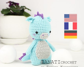 May include: A light blue crocheted dragon amigurumi toy with a purple crest, grey belly, and black eyes. The toy is standing on a white surface with white balls in the background. The text "TANATI CROCHET BY TATIANA KUCHEROVSKA" is at the bottom of the image. The image also includes flags for the United States, France, and Germany.