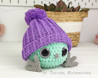May include: A crocheted green and purple toy with large eyes and a purple knitted hat. The toy is sitting on a white wooden surface. The text "TANATICROCHET BY TATIANA KUCHEROVSKA" is visible below the toy.