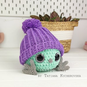 Pode incluir: Um brinquedo de crochê verde e roxo com olhos grandes e um chapéu de tricô roxo. O brinquedo está sentado em uma superfície de madeira branca. O texto "TANATICROCHET BY TATIANA KUCHEROVSKA" é visível abaixo do brinquedo.