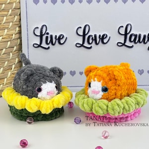 Może przedstawiać: Dwie ręcznie robione zabawki dla kotów na szydełku. Jedna jest szara i biała, druga pomarańczowa i biała. Obie siedzą w kolorowej podstawie w kształcie kwiatu. Tło zawiera słowa "Live Love Laugh".