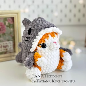 Puede incluir: Un gato de crochet con un disfraz de tiburón gris. El gato es blanco con manchas naranjas y tiene ojos negros. El disfraz de tiburón tiene una boca blanca con dientes negros.