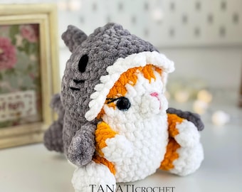May include: A crocheted cat wearing a gray shark costume. The cat is white with orange patches and has black eyes. The shark costume has a white mouth with black teeth.