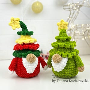 May include: Two handmade Christmas tree gnomes. One is red, yellow, and green with a yellow star on top. The other is green with a yellow star. Both have white beards and tan noses. The gnomes are on a white surface with Christmas lights in the background.