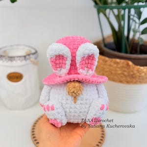 May include: A handmade crochet bunny gnome in white, pink, and beige. The gnome wears a pink hat with white-lined ears and has pink paws. The text "TANATIcrochet by Tatiana Kucherovska" is visible.