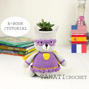 May include: Crochet superhero planter pattern in English, Spanish, and French. The pattern features a gray and purple superhero with a yellow cape and mask. The planter is holding a small succulent plant.