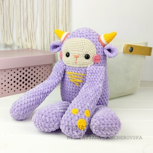 Könnte beinhalten: Ein gehäkeltes lila Monster-Spielzeug mit gelben Hörnern, orange-weißen Streifen und gelben Punkten. Das Spielzeug hat ein cremefarbenes Gesicht mit schwarzen Augen und rosa Wangen. Das Spielzeug sitzt auf einer weißen Oberfläche. "TANATi CROCHET BY TATIANA KUCHEROVSKA" steht auf der Oberfläche.