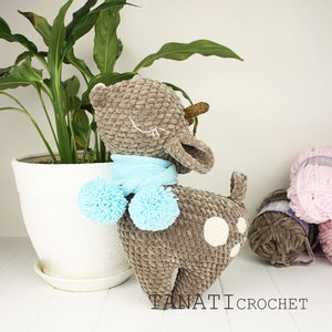 May include: A crocheted fawn with a blue scarf and pom poms. The fawn is brown with white spots and has closed eyes. The fawn is standing on a white surface.