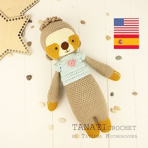 Puede incluir: Un juguete de perezoso tejido a ganchillo con pelaje marrón claro, cara color crema y parches oculares de color amarillo oscuro. Lleva una camisa azul claro con un pequeño corazón rosa. El texto TANATICROCHET BY TATIANA KUCHEROVSKA es visible.