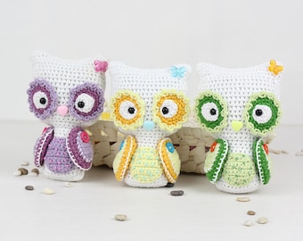 May include: Three crocheted owl toys, each with a different colored body and eyes. The owls are white with purple, yellow, and green accents. The owls are sitting on a white surface.