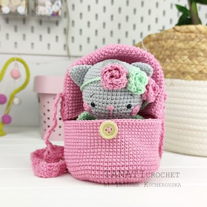 May include: A crocheted pink bag with a gray crocheted cat inside. The cat has pink and green flowers on its head. The bag has a strap and a yellow button.
