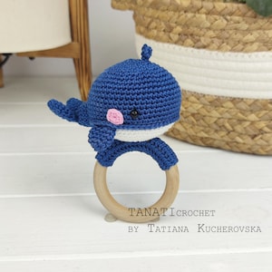 May include: Blue crocheted whale rattle with a wooden ring. The whale has a pink nose and black eyes. The text "TANATI CROCHET BY TATIANA KUCHEROVSKA" is printed below the rattle.