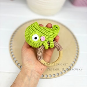 May include: Green crocheted chameleon rattle toy with a wooden ring. The chameleon has a pink nose and a black eye. The text "TANATI CROCHET BY TATIANA KUCHEROVSKA" is visible on the wooden ring.