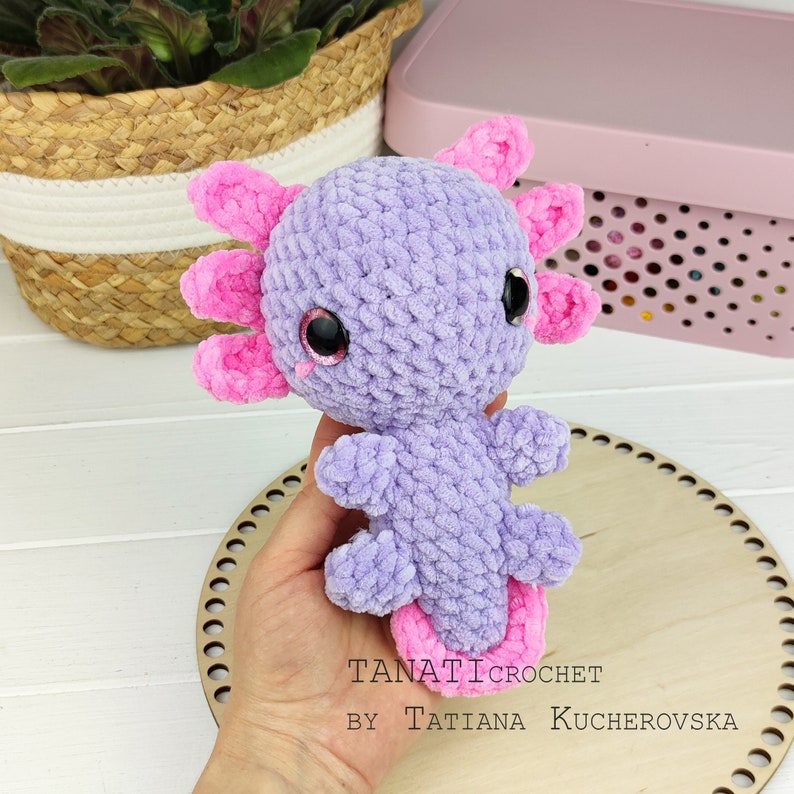 May include: A crocheted axolotl plush toy with pink gills and purple body. The toy has large black eyes with pink accents. The toy is held in a hand against a wooden background.
