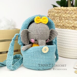 Pode incluir: Um brinquedo de elefante de croch&ecirc; com um la&ccedil;o amarelo est&aacute; sentado dentro de uma bolsa de croch&ecirc; azul com fecho de bot&atilde;o. A bolsa tem uma al&ccedil;a longa, o que a torna uma bolsa de ombro.