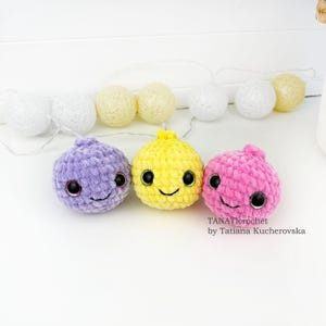 May include: Three round, plush, crocheted toys in purple, yellow, and pink, each featuring large black safety eyes and a stitched smile. Behind them is a string of white and pale yellow decorative globe lights. Text reads TANATIcrochet by Tatiana Kucherovska.