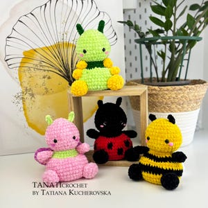 Könnte beinhalten: Eine Sammlung handgefertigter Häkel-Insekten-Plüschtiere. Die Spielzeuge umfassen eine grüne Raupe, einen rosa und lila Käfer, einen schwarzen Marienkäfer mit roten Punkten und eine gelb-schwarze Biene. Die Spielzeuge werden auf einer weißen Oberfläche präsentiert.
