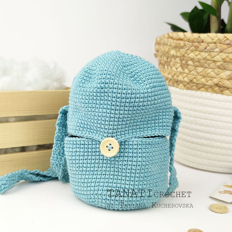 Pode incluir: Um chap&eacute;u de balde de croch&ecirc; azul claro com fecho de bot&atilde;o e la&ccedil;os compridos. O chap&eacute;u est&aacute; dobrado para baixo e os la&ccedil;os est&atilde;o amarrados juntos. O texto "TANATI CROCHET by TATIANA KUCHEROVSKA" &eacute; vis&iacute;vel na parte inferior do chap&eacute;u.