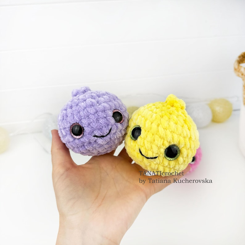 May include: Two round, plush, crocheted toys with soft, fuzzy yarn textures. One toy is light purple and the other is bright yellow. Both feature large, dark, glossy safety eyes and a simple stitched smile. The text TANATIcrochet by Tatiana Kucherovska is visible.