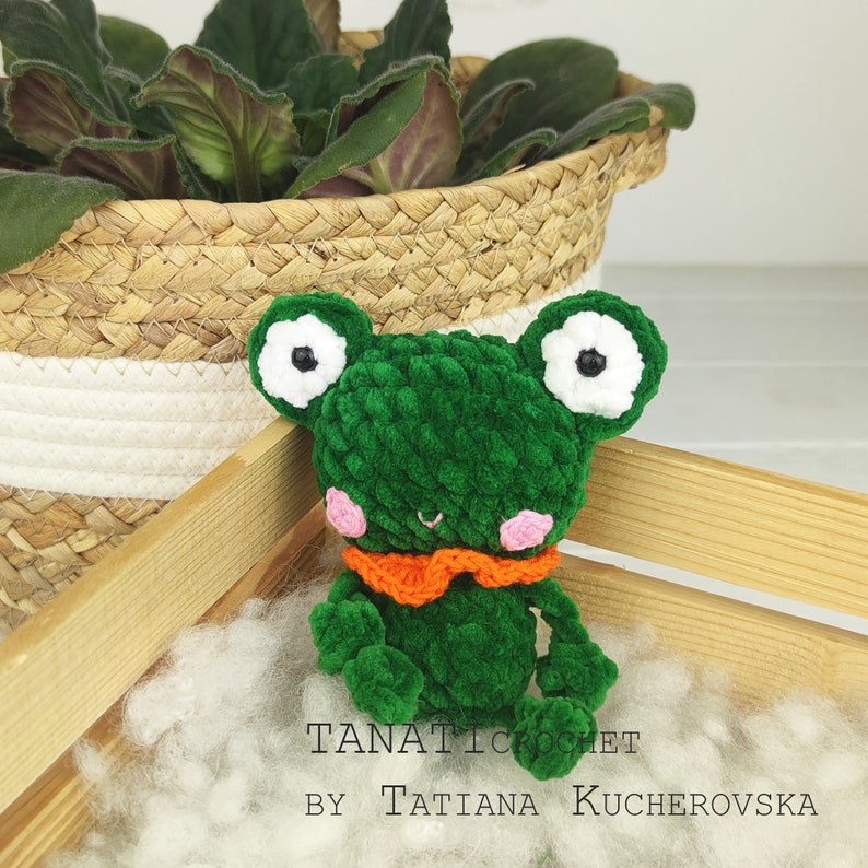 May include: Green crocheted frog toy with white eyes, pink cheeks, and an orange bow tie. The frog is sitting on a white fluffy surface.