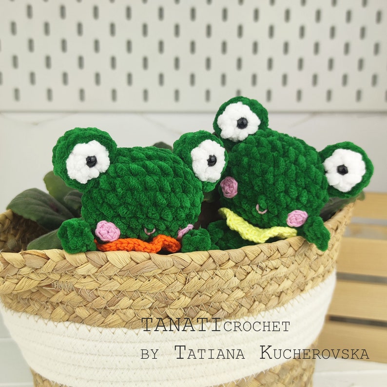 May include: Two green crocheted frog plushies with white eyes and pink cheeks, sitting in a woven basket. The basket has a white stripe around the top. The text "TANATI CROCHET BY TATIANA KUCHEROVSKA" is visible at the bottom of the image.