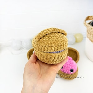 May include: A round, golden-brown crocheted container with a lid, featuring a textured yarn surface. A small pink crocheted creature with black eyes is visible nearby. The text TANATicrochet by Tatiana Kucherovska is printed on the image.
