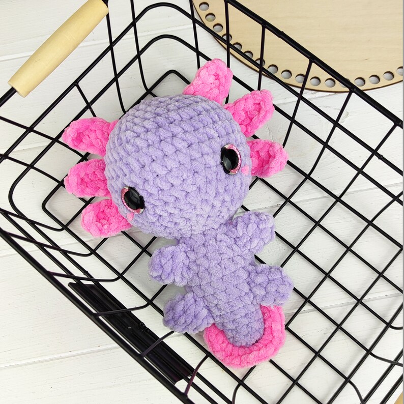 May include: A purple and pink crocheted axolotl plush toy with large black eyes. The toy is sitting in a black wire basket.