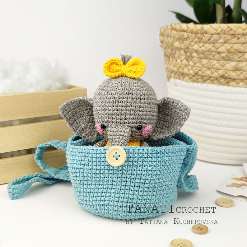 Pode incluir: Um elefante de croch&ecirc; cinza com um la&ccedil;o amarelo est&aacute; sentado dentro de uma cesta de croch&ecirc; azul claro com um bot&atilde;o branco. A cesta tem uma al&ccedil;a longa.