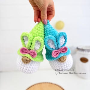 K&ouml;nnte beinhalten: Zwei handgefertigte H&auml;kelzwerg-Ornamente. Eines ist gr&uuml;n und das andere blau, beide mit wei&szlig;en Ohren, rosa Schleifen und braunen Nasen. Die Zwerge haben wei&szlig;e K&ouml;rper und werden von einer Hand gehalten.