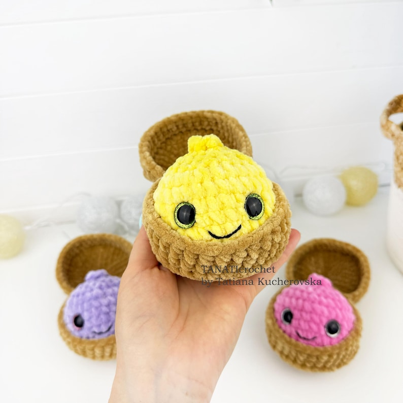 May include: A yellow crocheted plush toy with black eyes and a stitched smile, nestled inside a brown crocheted shell. The text TANATcrochet by Tatiana Kucherovska is visible on the yellow plush. Two additional plush toys in purple and pink are nearby.