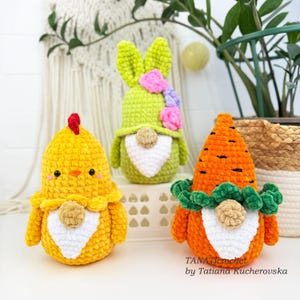 K&ouml;nnte beinhalten: Drei handgefertigte H&auml;kelfiguren zu Ostern: ein gelbes K&uuml;ken, ein gr&uuml;nes H&auml;schen mit rosa und lila Blumen und eine orangefarbene Karotte mit gr&uuml;nen Bl&auml;ttern. Jede Figur hat einen wei&szlig;en Bart und eine neutralfarbene Nase.