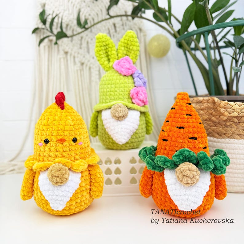 K&ouml;nnte beinhalten: Drei handgefertigte H&auml;kelfiguren zu Ostern: ein gelbes K&uuml;ken, ein gr&uuml;nes H&auml;schen mit rosa und lila Blumen und eine orangefarbene Karotte mit gr&uuml;nen Bl&auml;ttern. Jede Figur hat einen wei&szlig;en Bart und eine neutralfarbene Nase.
