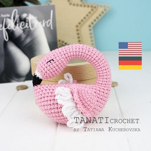 May include: Pink crocheted flamingo toy shaped like a ring. The toy has a white crocheted wing and black eyes. The toy is on a wooden surface. The text "TANATI CROCHET BY TATIANA KUCHEROVSKA" is below the toy.