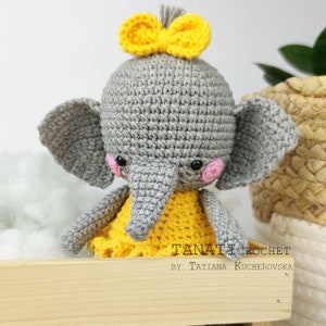 Pode incluir: Um elefante de croch&ecirc; cinza vestindo um vestido amarelo com um la&ccedil;o amarelo na cabe&ccedil;a. O elefante tem bochechas cor-de-rosa e olhos pretos. O texto "TANATICROCHET BY TATIANA KUCHEROVSKA" &eacute; vis&iacute;vel no canto inferior direito da imagem.