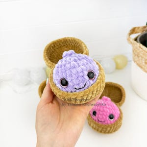 May include: A crochet plush toy featuring a light purple round character with large black eyes and a small smile, nestled inside a tan textured bowl. A second similar plush toy with a pink top is visible nearby. Text reads TANATicrochet by Tatiana Kucherovska.