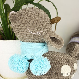 May include: A gray crocheted deer with a blue scarf and pom-poms. The deer has white spots and closed eyes. The deer is sitting on a white surface. The text "TANATI CROCHET" is visible on the bottom right of the image.