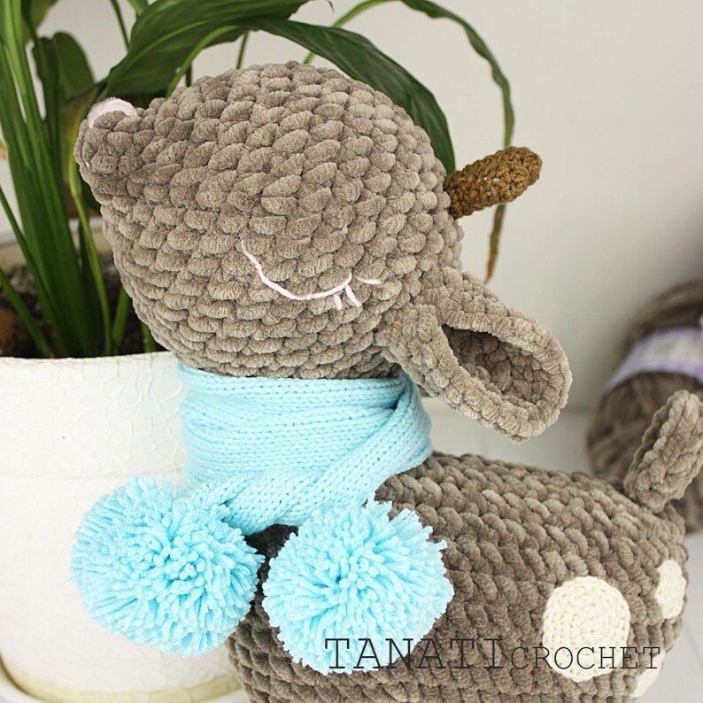 May include: A gray crocheted deer with a blue scarf and pom-poms. The deer has white spots and closed eyes. The deer is sitting on a white surface. The text "TANATI CROCHET" is visible on the bottom right of the image.