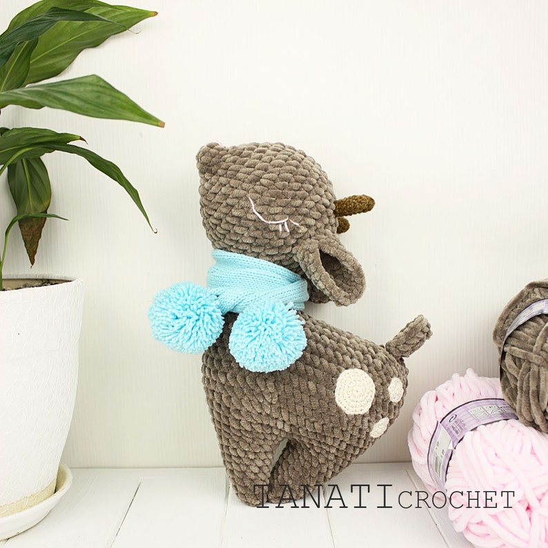 May include: A brown crocheted deer with a blue scarf and pom-poms. The deer has white embroidered eyes and a white spot on its back. The deer is standing on a white surface. The text "TANATI CROCHET" is visible in the bottom right corner of the image.