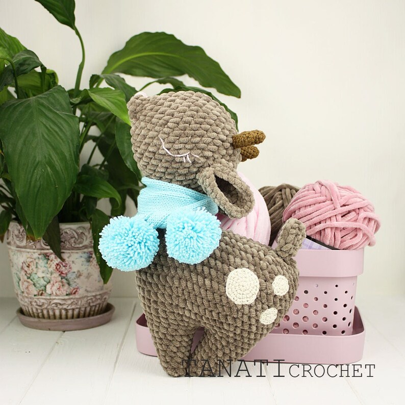 May include: A brown crocheted deer with a blue scarf and pom poms. The deer is sitting on a pink basket with yarn balls.