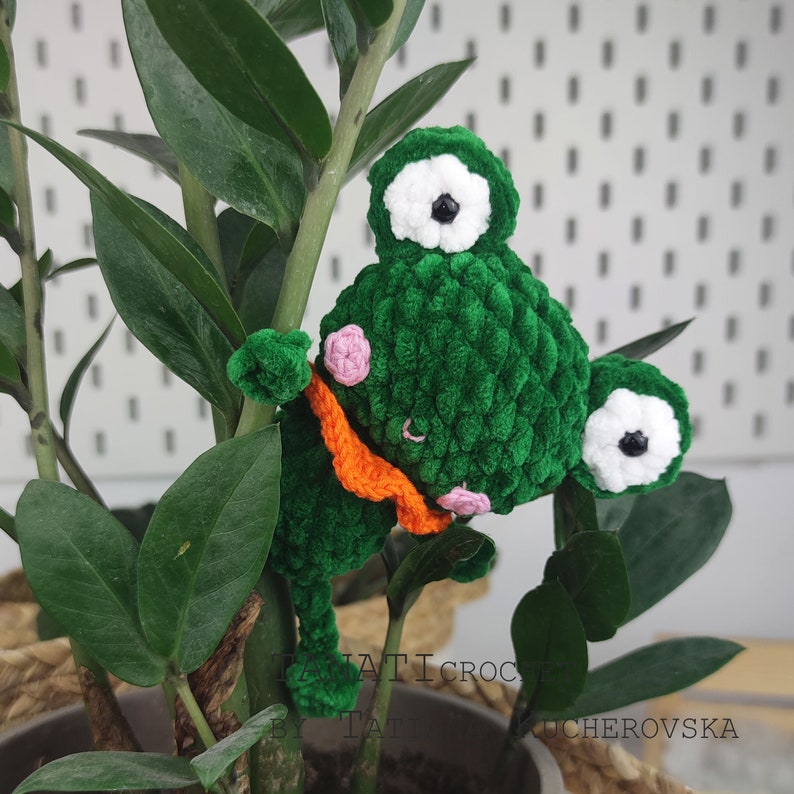 May include: A green crocheted frog with white eyes and an orange scarf. The frog is hanging on a green plant.